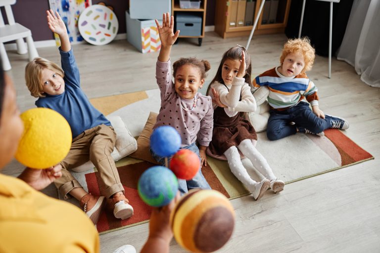 Adorable little girl raising hand while sitting on carpet between classmates